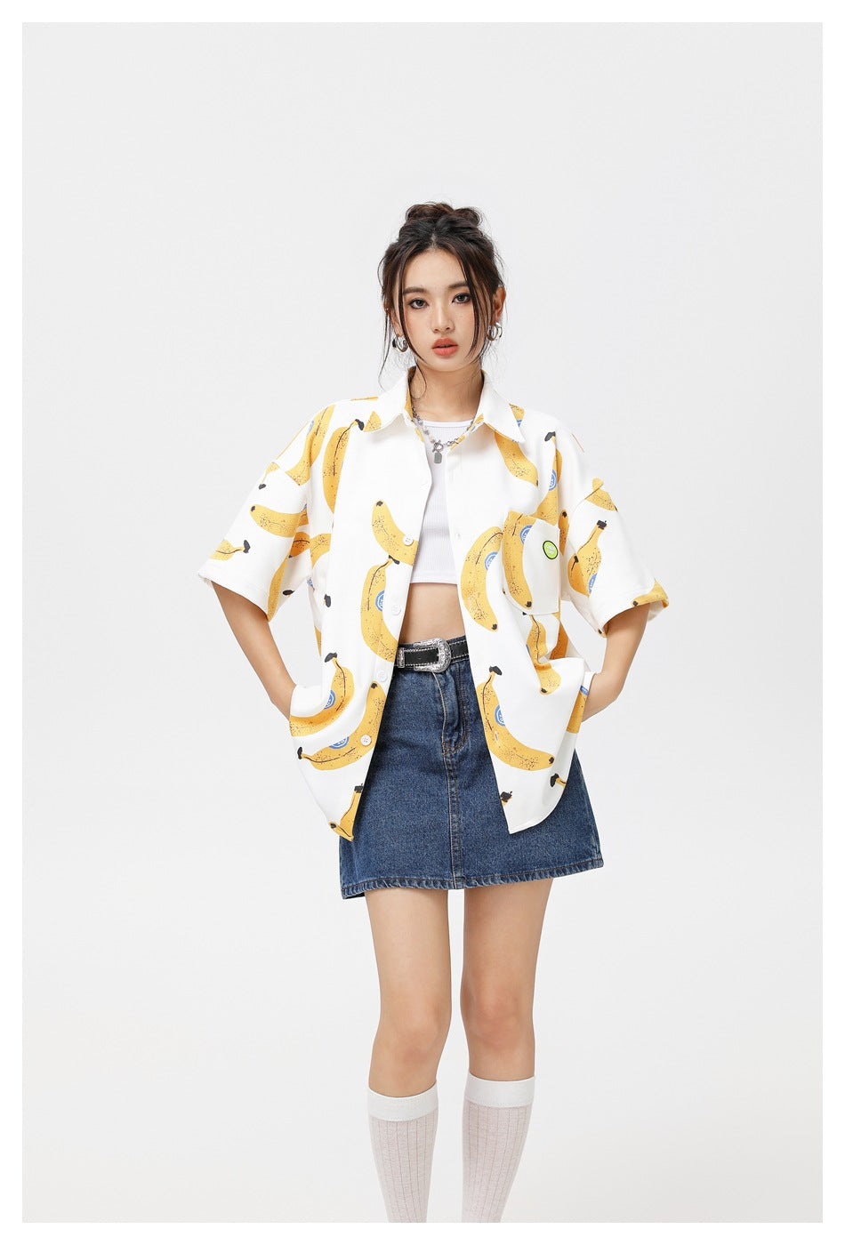 Model wearing a white short-sleeve banana-print shirt open over a white crop top with a denim mini skirt