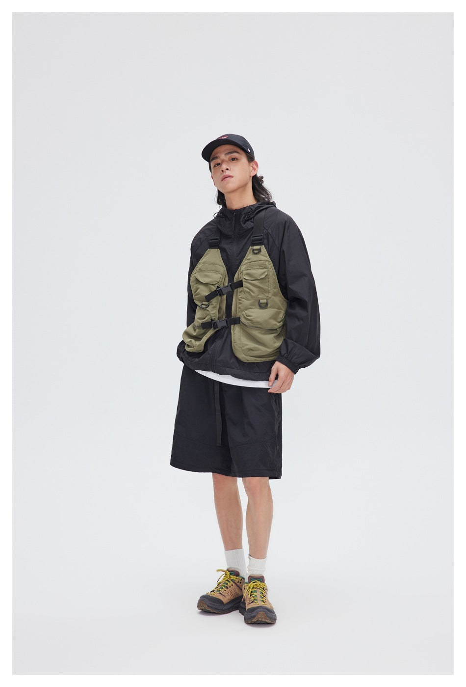Model wearing a black hooded jacket, olive utility vest, and black shorts.