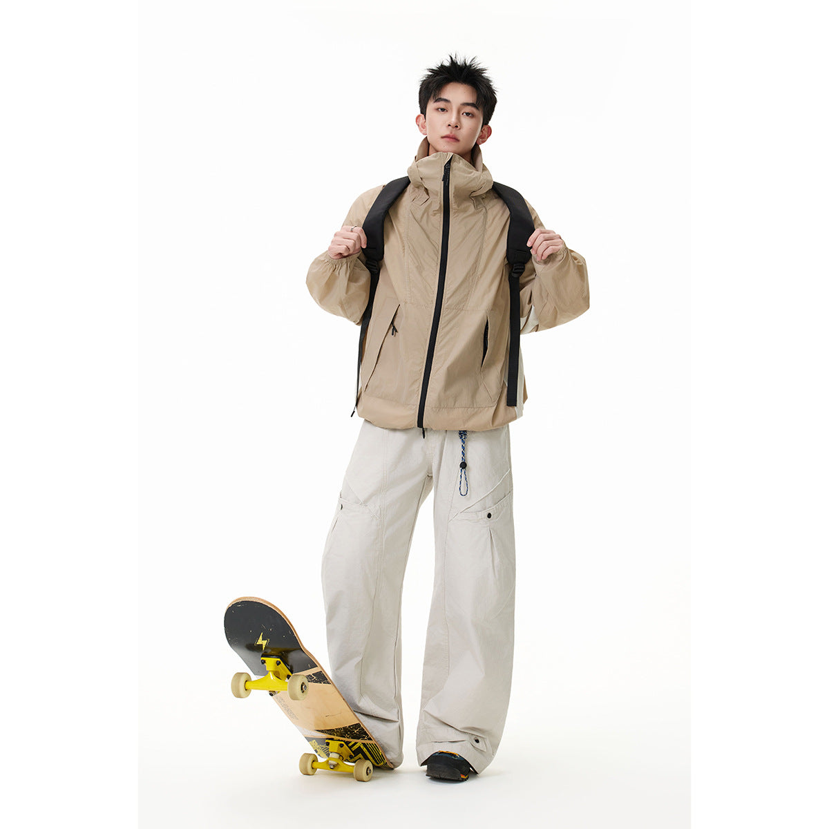 Full-length model wearing a tan high-collar zip jacket and wide light trousers, holding a skateboard against a white