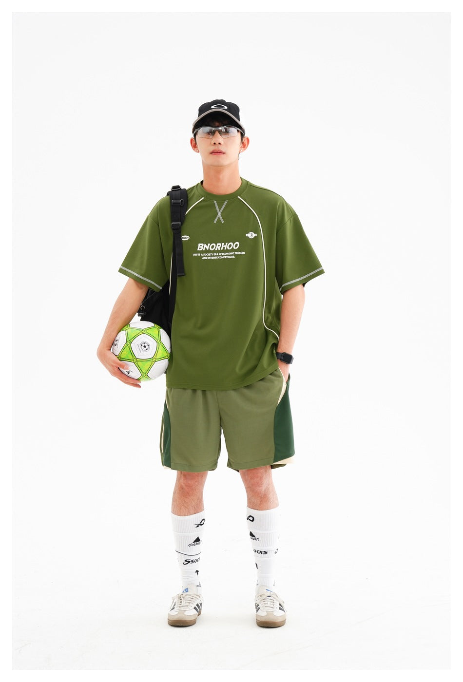 Model wearing an olive green athletic T-shirt and color-block shorts, carrying a soccer ball in a shoulder bag.