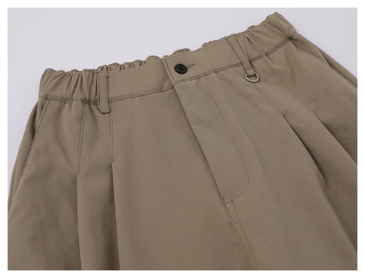 Close-up of taupe trousers showing the gathered waistband, button, belt loop, metal ring, and front pleats.
