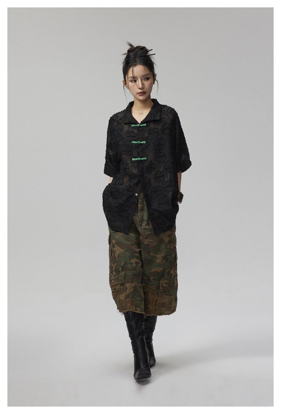 Model walking in black floral-textured short-sleeve shirt with green knot closures, camouflage cargo shorts and black boots