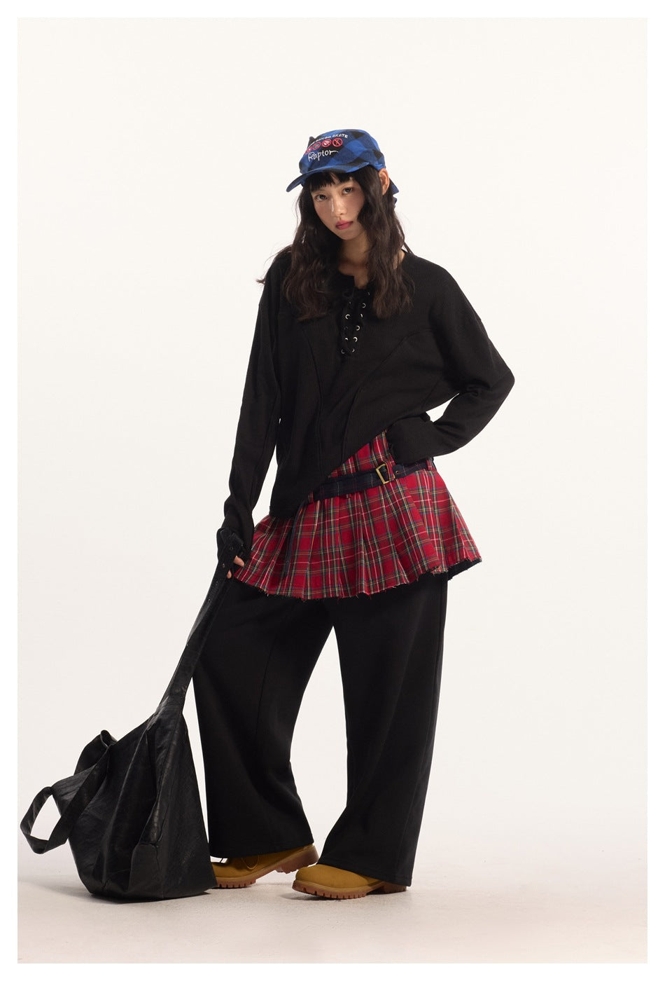 Model posing in black lace-up long-sleeve top over red plaid pleated skirt and wide black pants with yellow boots