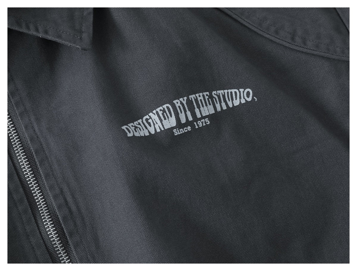 Close-up of a charcoal jacket with a light printed “Designed by the Studio, Since 1975” chest graphic and zipper.