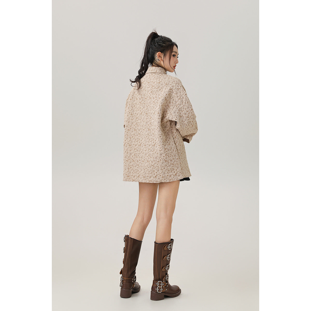 Rear view of a model wearing an oversized beige floral jacket with brown knee-high boots.