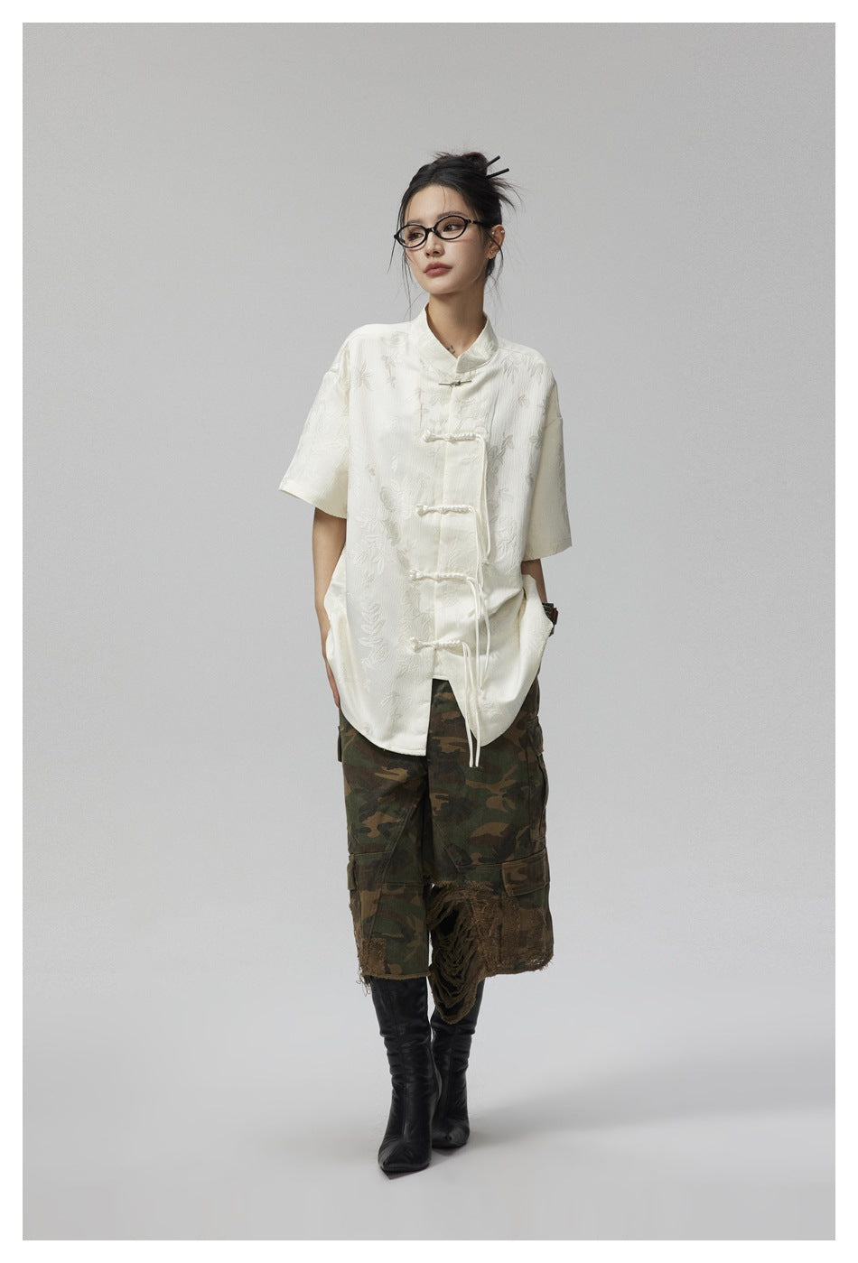 Woman wearing an off-white short-sleeve frog-closure shirt with camo cargo shorts and black boots, front view