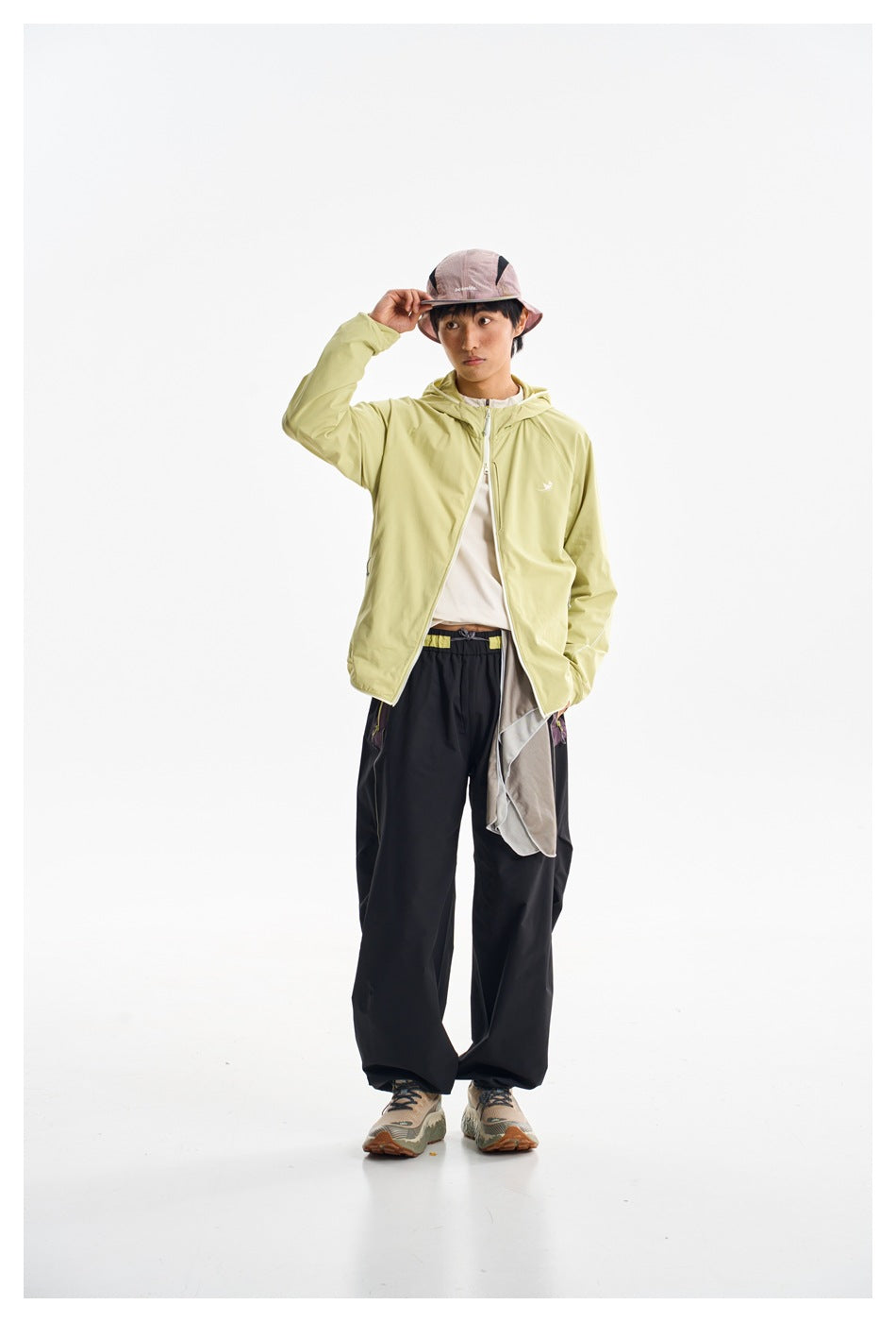 Model wearing a pale yellow-green hooded zip jacket with black trousers, a pink bucket hat and trail shoes.