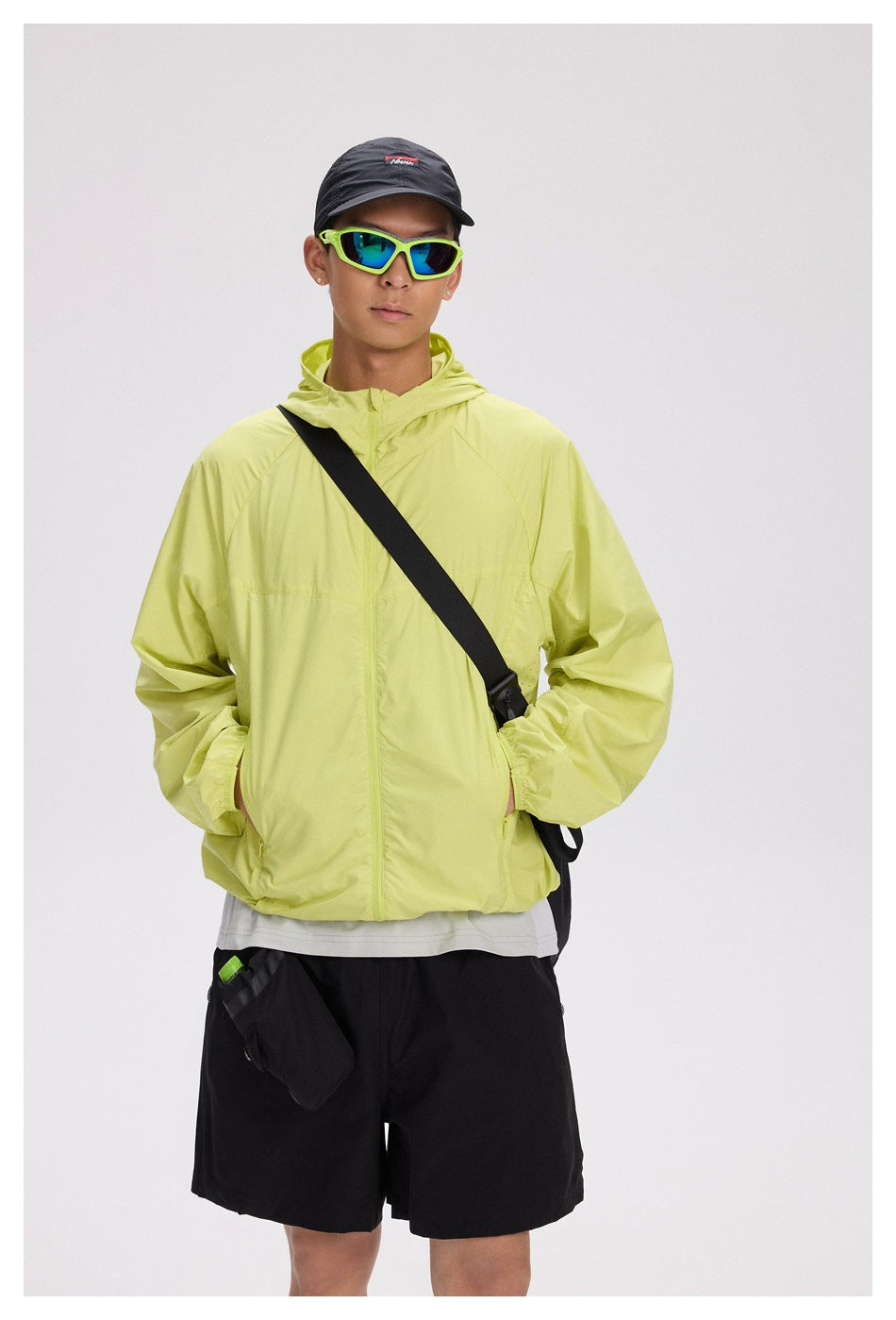 A model wears a bright lime hooded jacket with black shorts, a black cap, and neon green sunglasses.