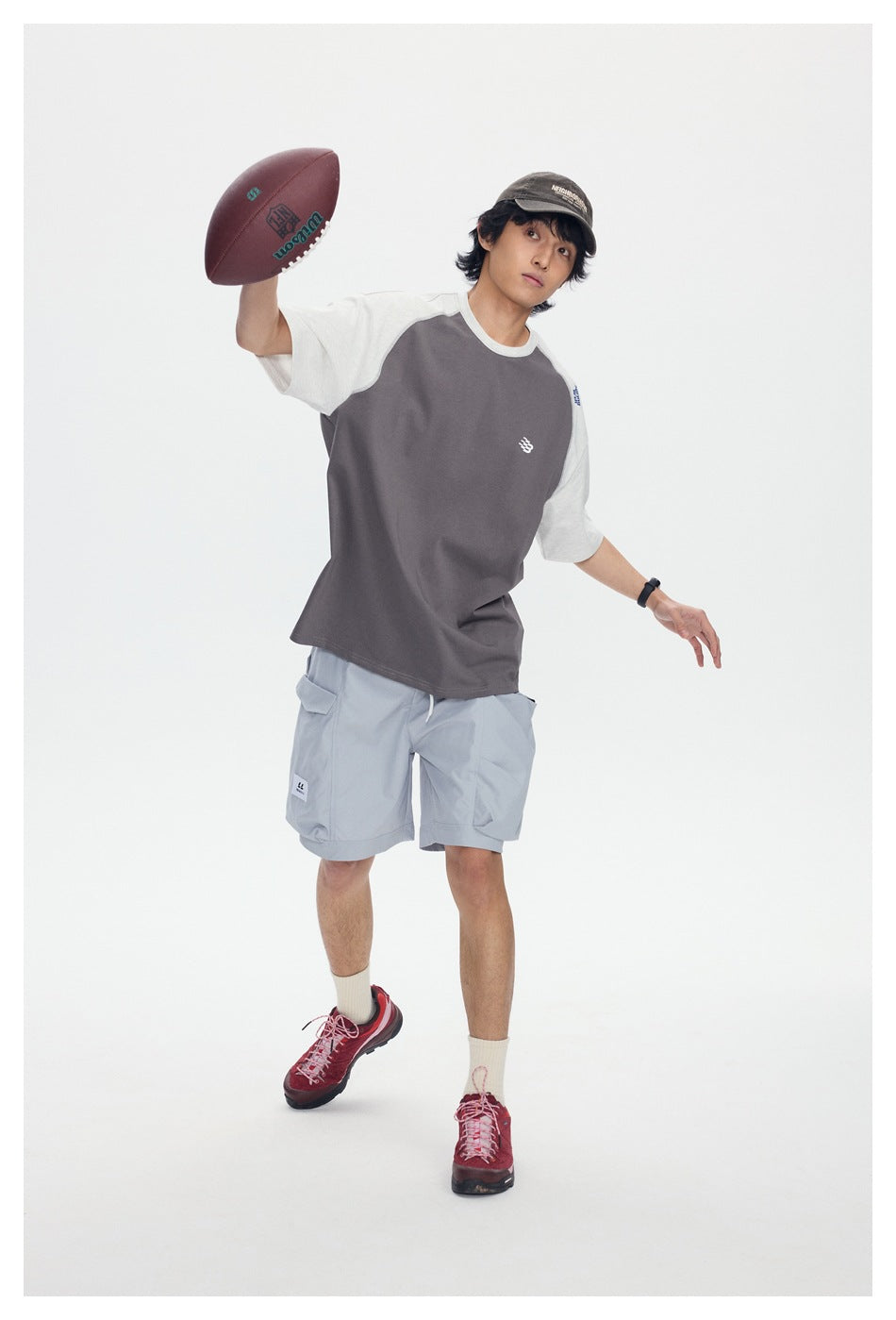 Model wearing a charcoal and light gray color-block raglan T-shirt with gray cargo shorts, holding a football.