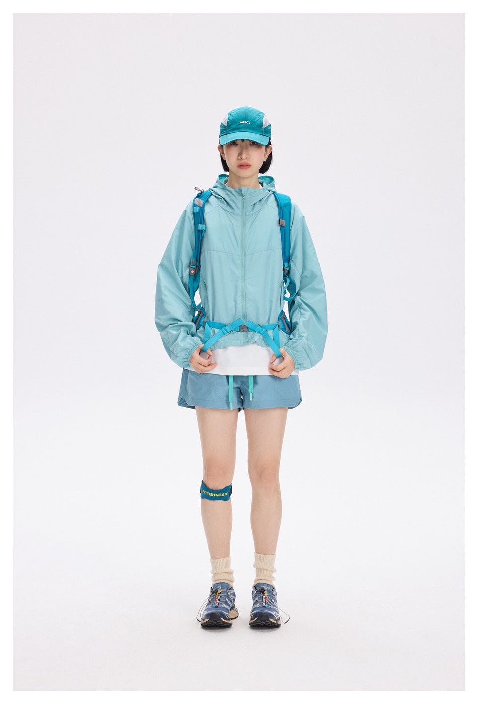Front view of a model wearing a light turquoise hooded jacket and shorts with a blue hiking backpack.