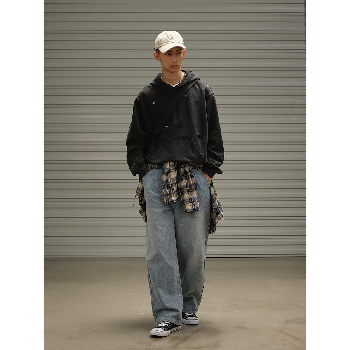 Model wearing a black distressed hoodie, loose blue jeans and cream cap, with a plaid shirt tied around the waist.