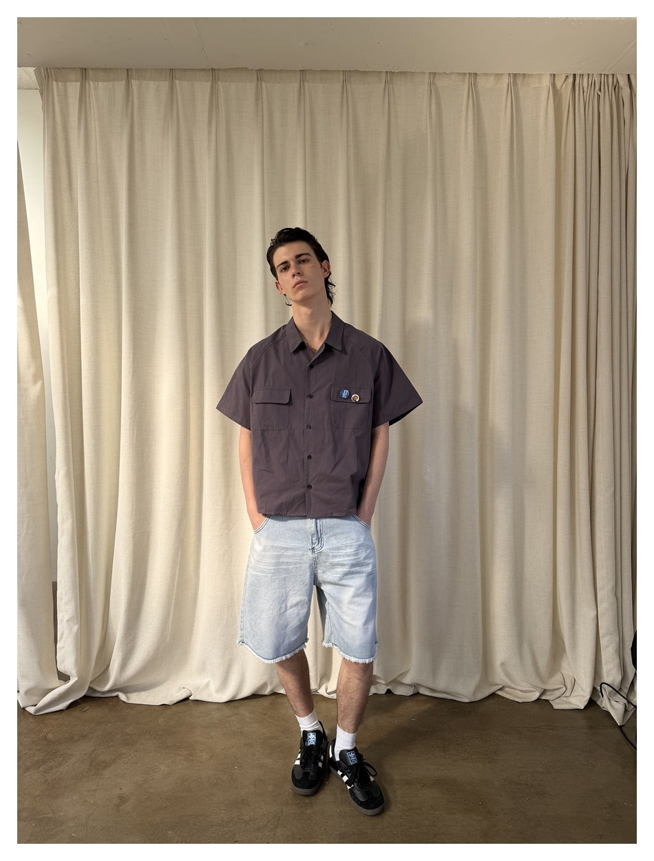 Model wearing a charcoal short-sleeve button-up shirt with two chest pockets and light wash frayed denim shorts, front view.