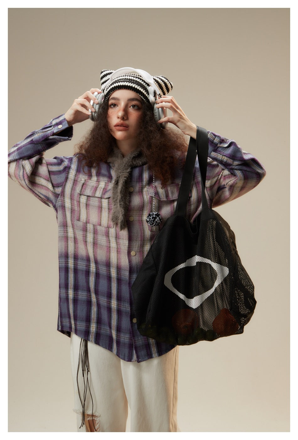 Close-up of a model wearing a purple plaid shirt jacket and striped beanie, holding headphones beside a mesh tote.