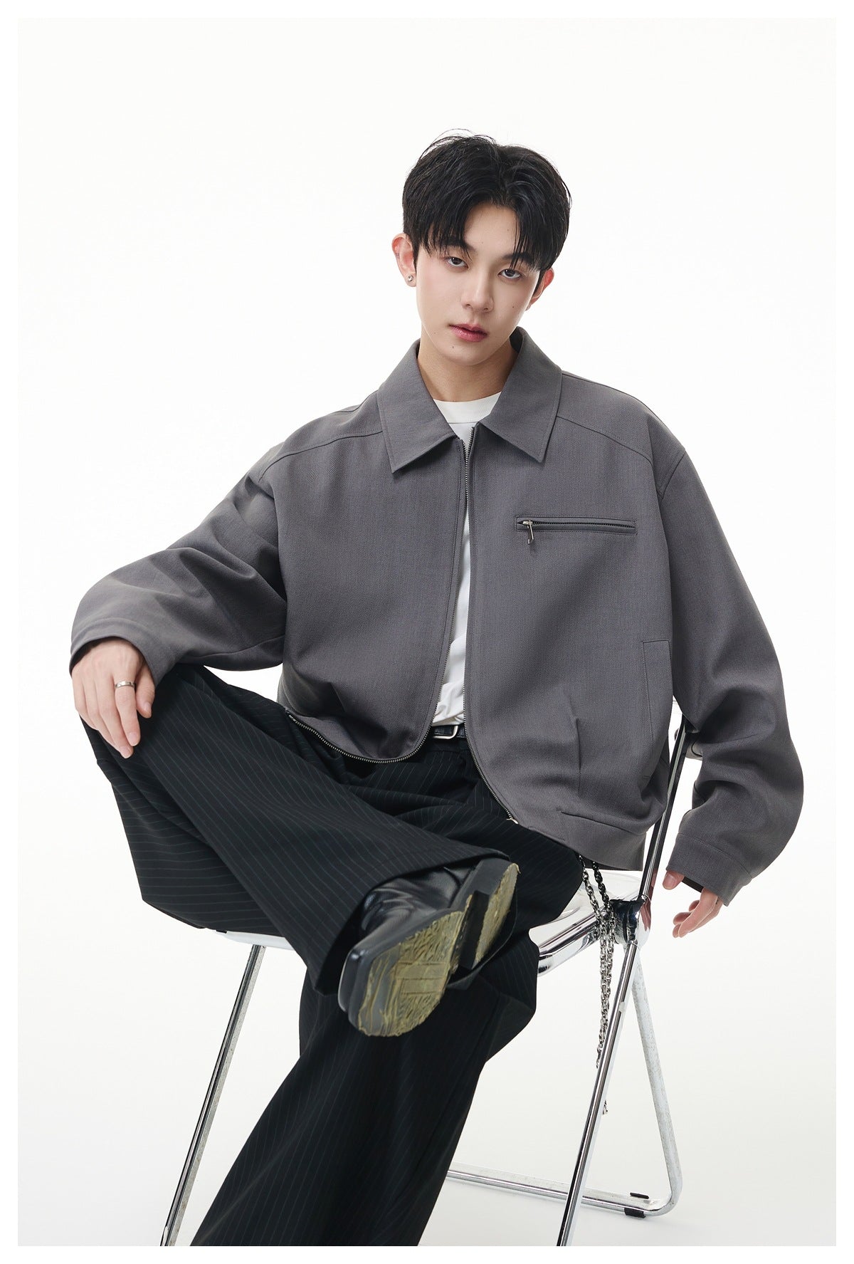 Model seated in a charcoal gray zip-front jacket with a pointed collar and chest zipper pocket.