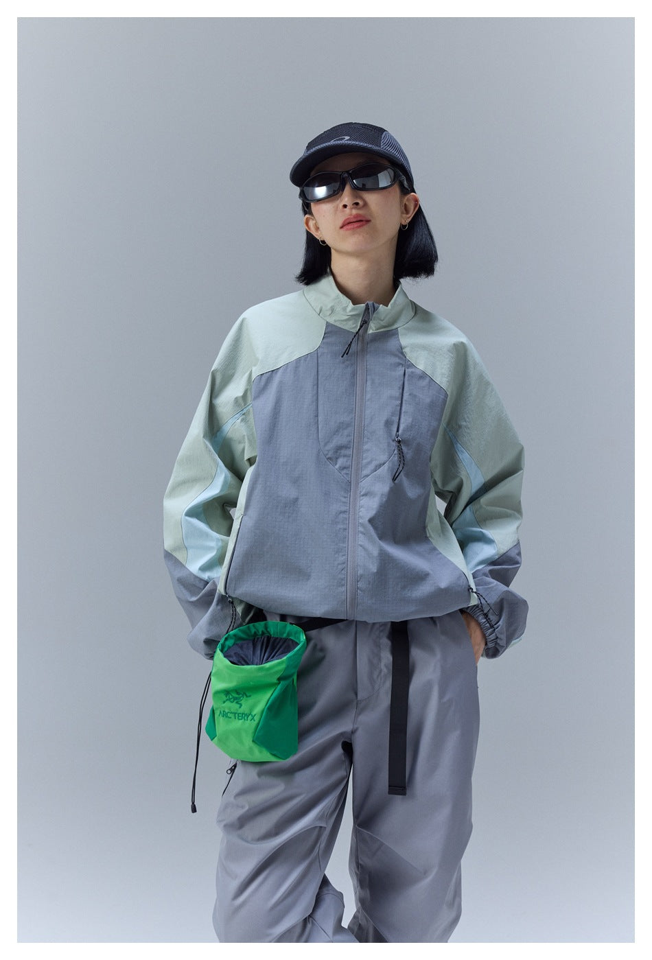 Front view of a model in a pale mint and blue-gray color-block jacket and gray pants, carrying a green pouch.
