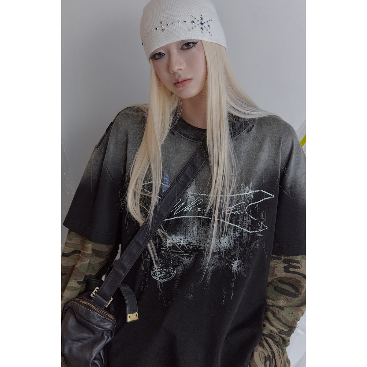 Model wearing an oversized black graphic tee over camouflage sleeves, with a white studded beanie and crossbody bag.