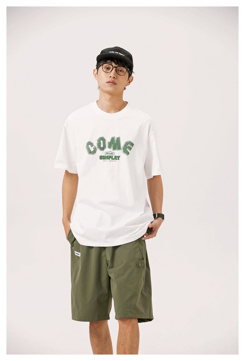 Model wearing white graphic T-shirt with green COME COMPLAY print, cap, glasses and olive green shorts