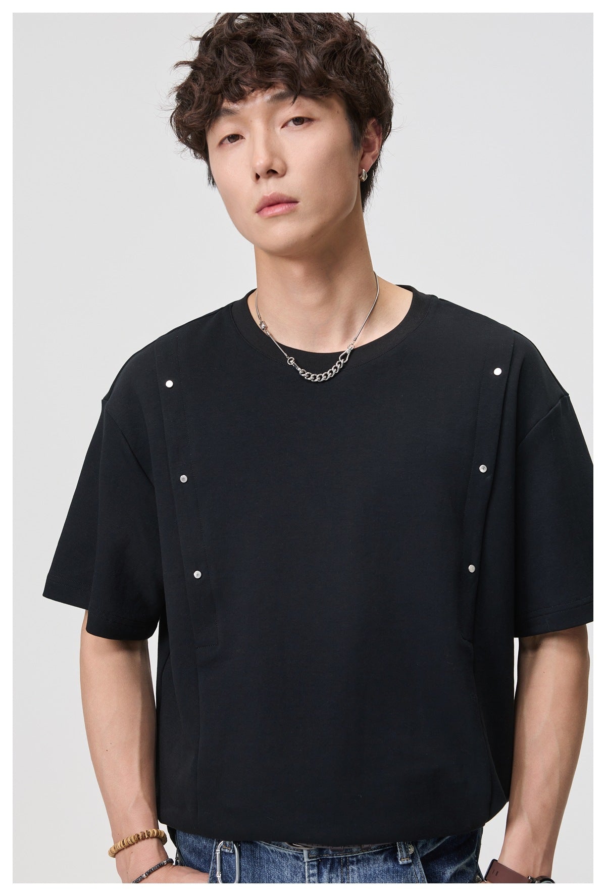 Man wearing a black crew-neck T-shirt with silver rivet studs on the front seams, close-up with chain necklace