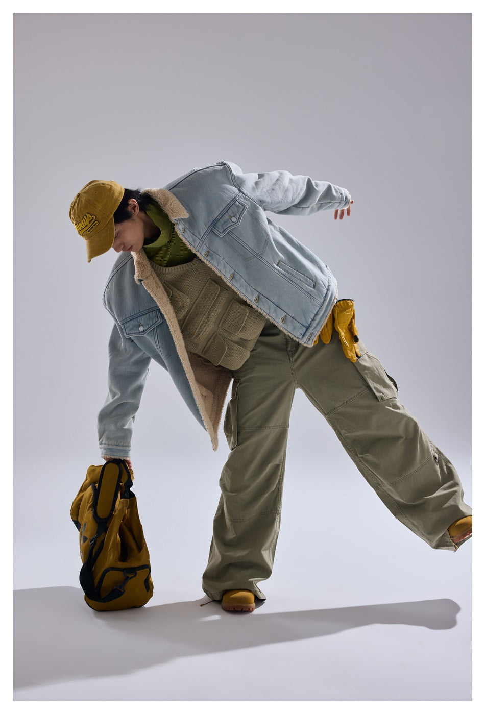 Model leans forward in a light blue sherpa-lined denim jacket and olive cargo pants, holding a yellow backpack.