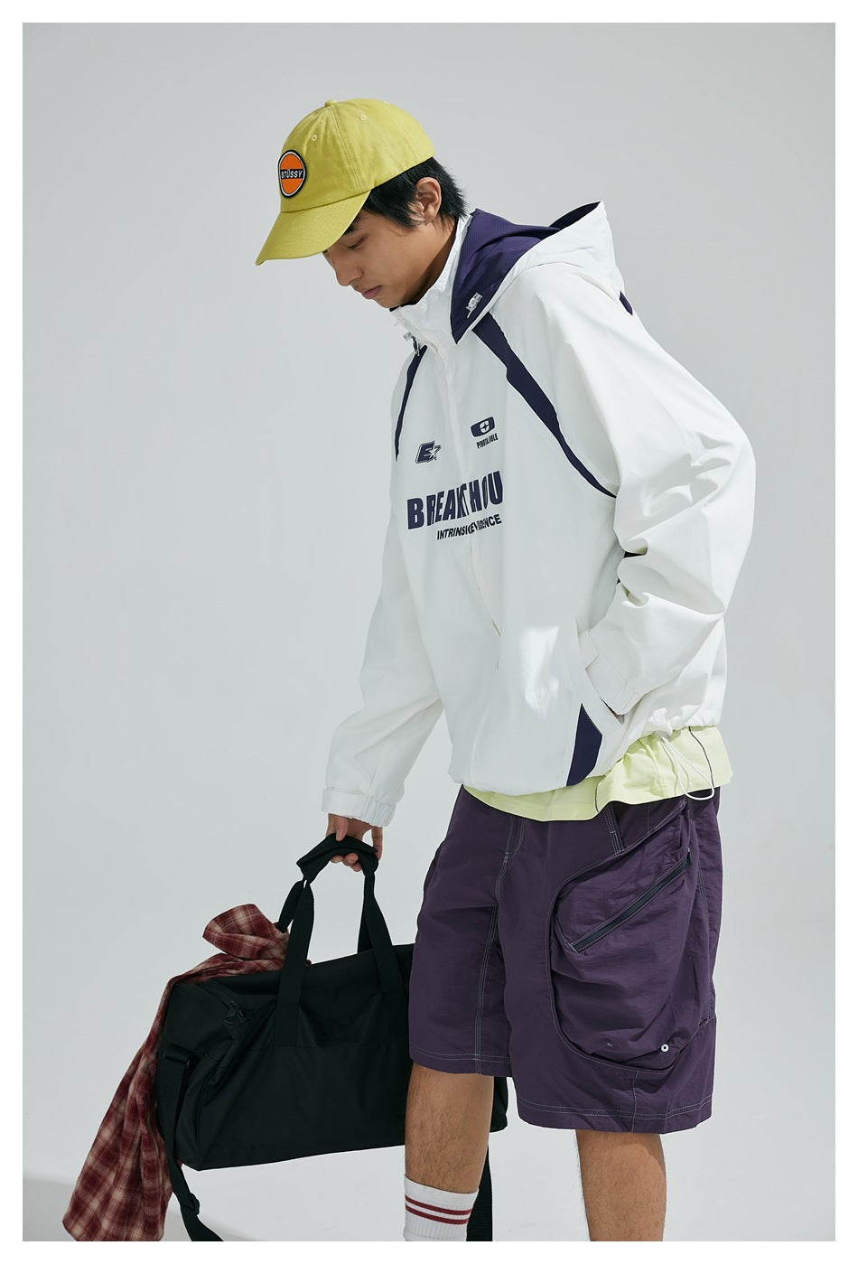 Model wearing a white hooded jacket with navy accents and chest graphics, purple shorts, and a yellow cap.