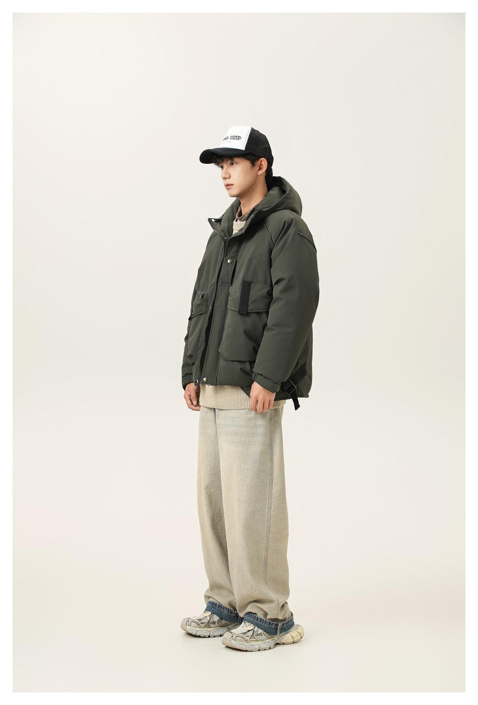 Model in army green hooded puffer jacket, side view, with beige wide-leg pants and a trucker cap