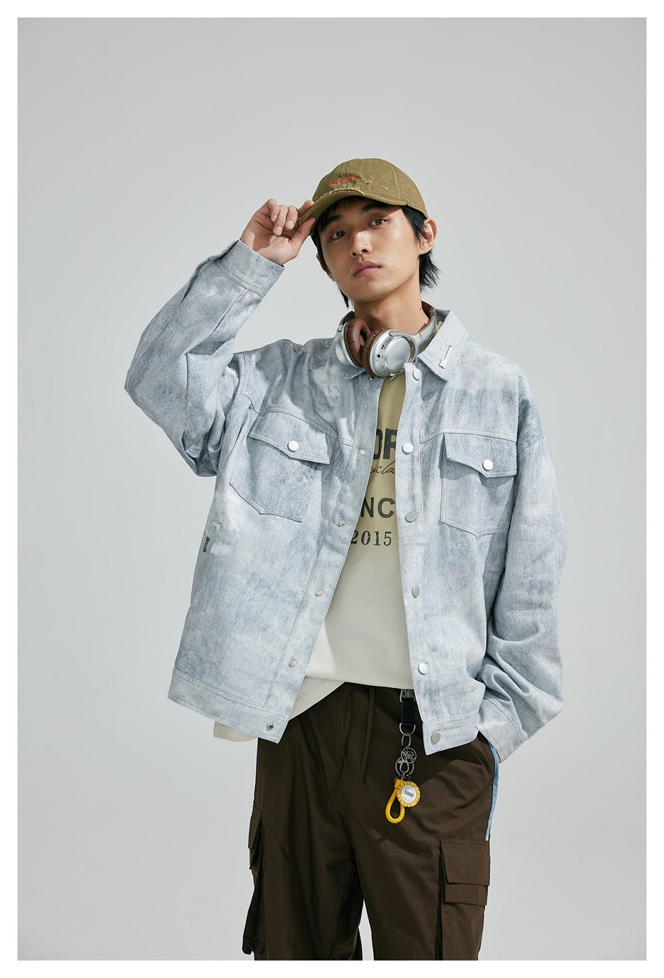 Model in light gray washed snap-front jacket over a cream tee and brown cargo pants, hand raised to cap