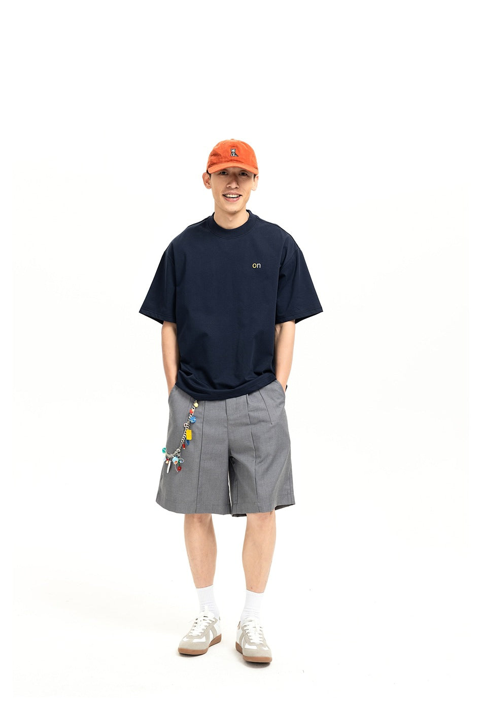 Man wearing navy T-shirt with small yellow 'on' chest print, gray shorts, orange cap and sneakers, front view