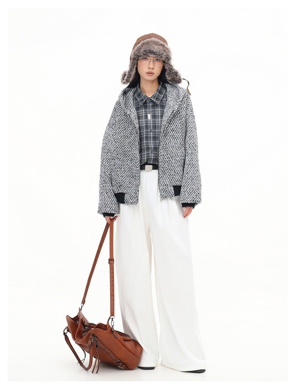 Full-length model in an open black-and-white textured hooded jacket, plaid shirt and white trousers, carrying a brown bag.