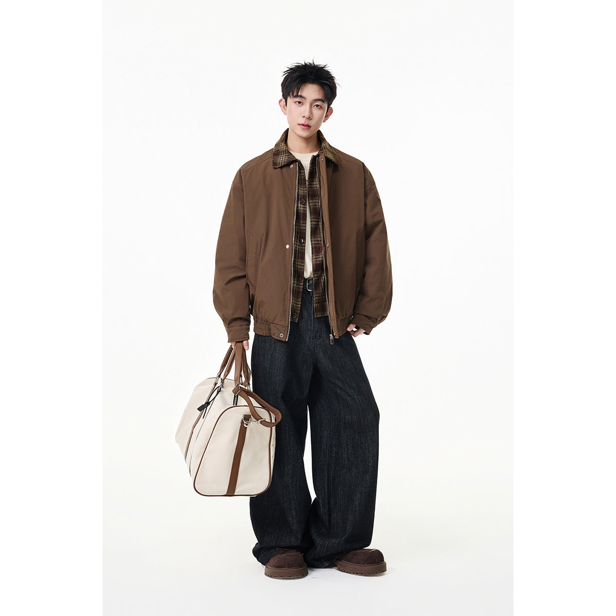 Full-length model wearing a brown jacket with a plaid collar, wide dark jeans, and carrying a cream duffel bag.