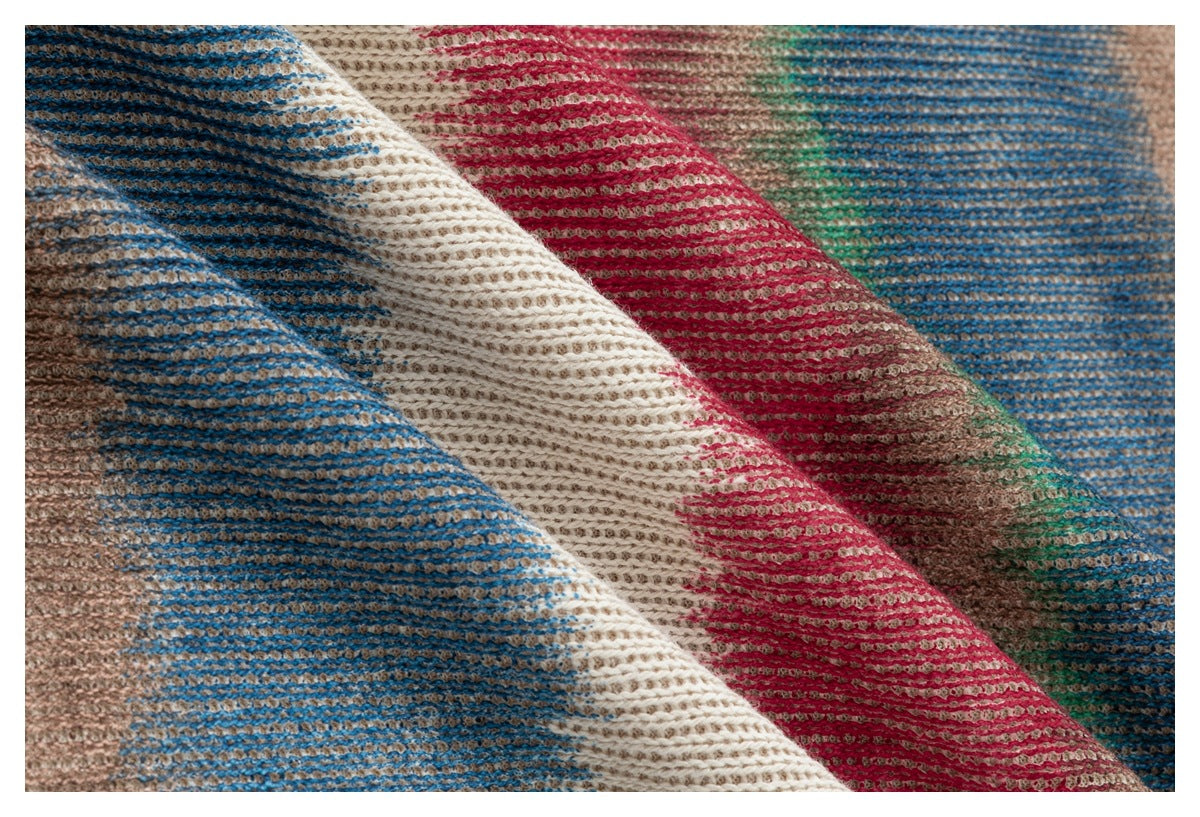 Extreme close-up of textured knit fabric with diagonal folds and bands of blue, cream, red, green, and tan.