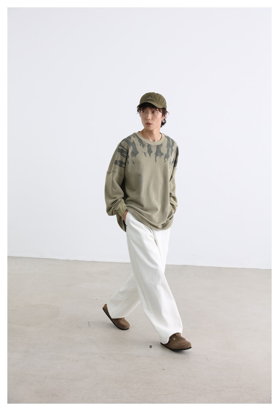 Full-length model wearing a khaki graphic sweatshirt, white wide-leg pants and an olive cap.