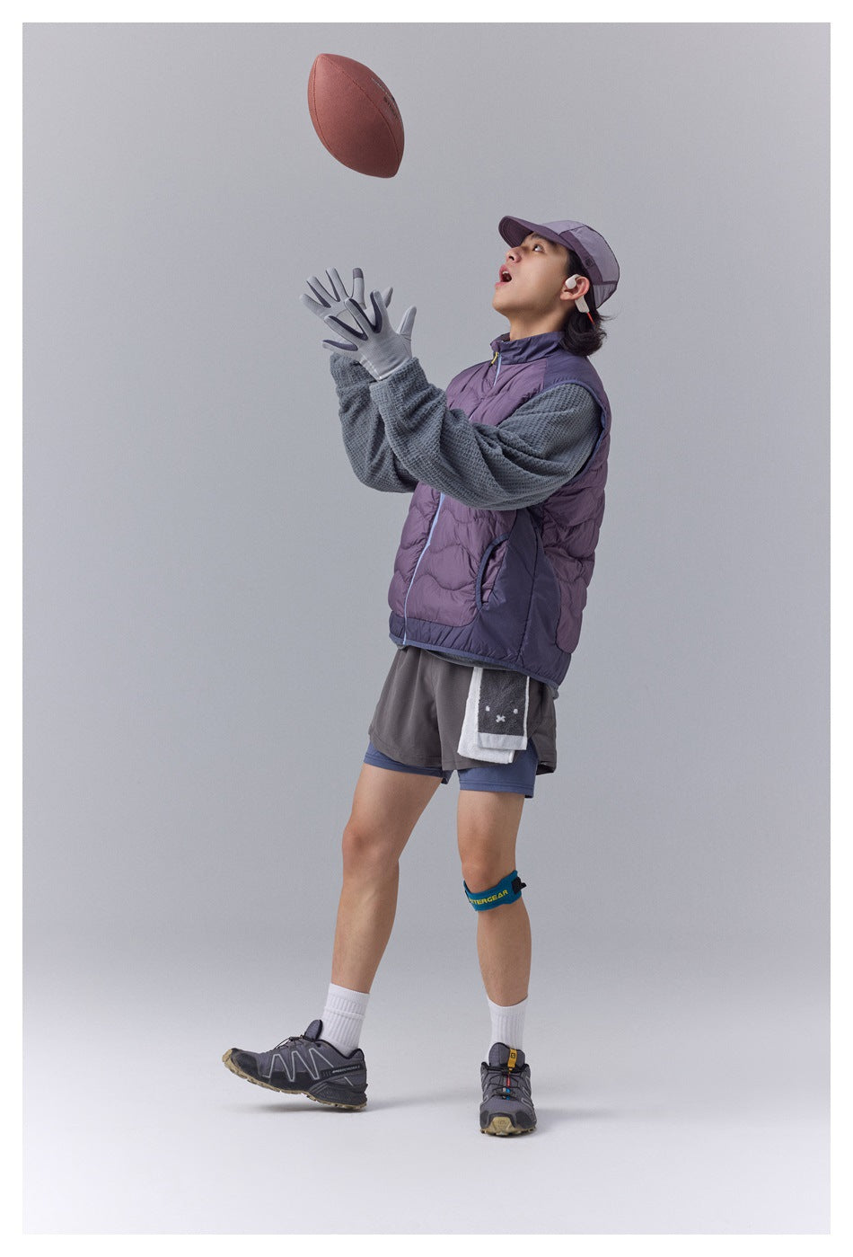 Model wearing a purple quilted vest over a textured gray pullover, catching a football; front view.