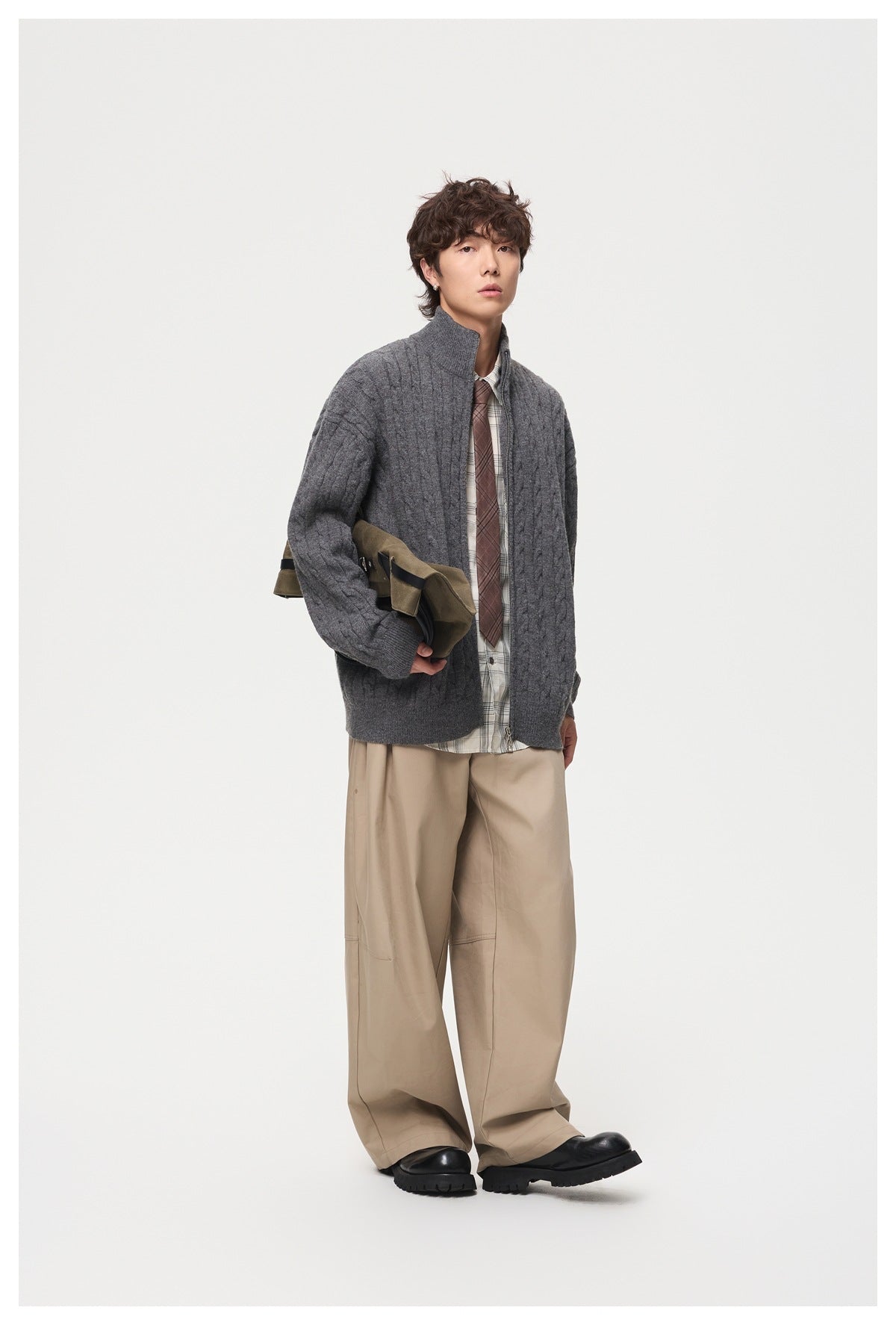 Model wearing a charcoal cable-knit cardigan with a high collar over a plaid shirt, tie, and beige trousers.