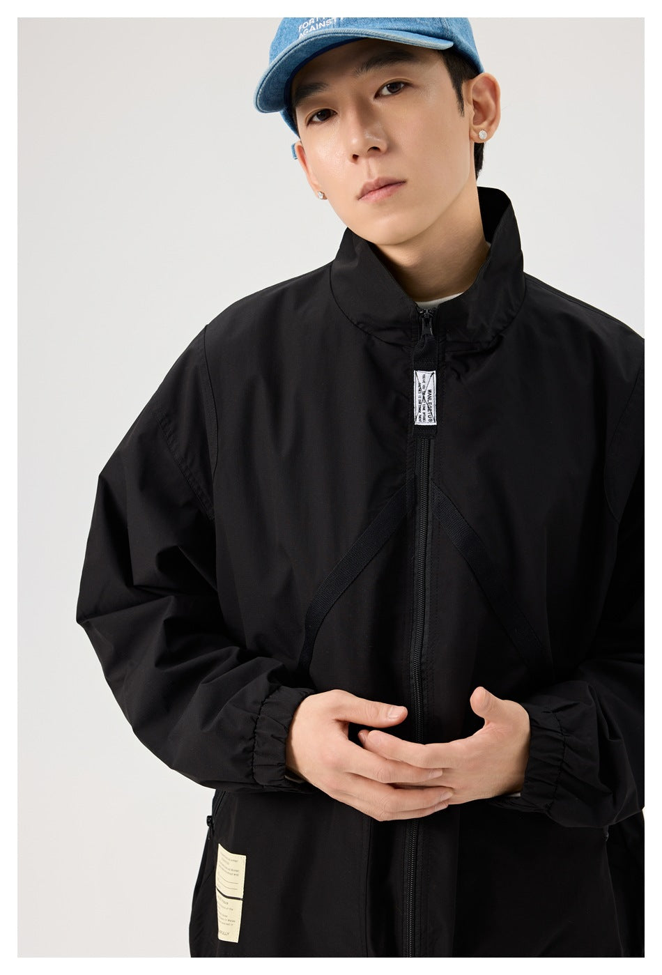 Model wearing a black zip-front jacket with a high collar and diagonal chest straps, paired with a blue cap.