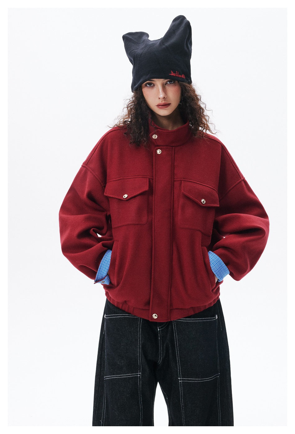 Closer front view of a model wearing a red jacket with a high collar, buttoned flap pockets and blue checked cuffs.