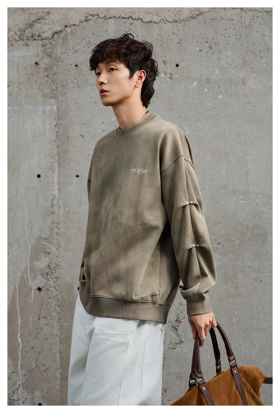 Model wearing an army green mottled crewneck sweatshirt with sleeve studs, white pants, and a brown bag against concrete