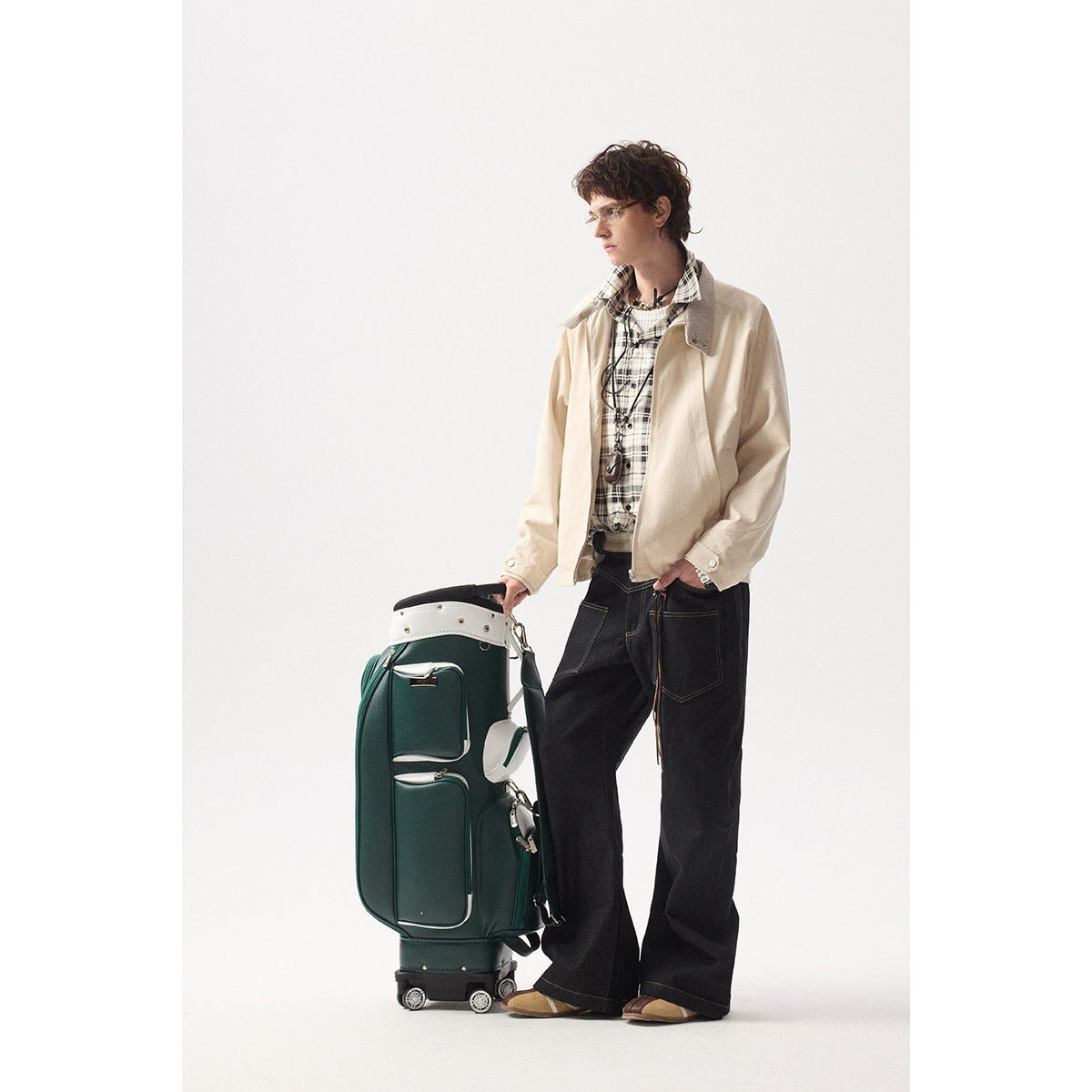 Model in a light beige jacket over a plaid shirt and dark jeans, standing beside a dark green suitcase.
