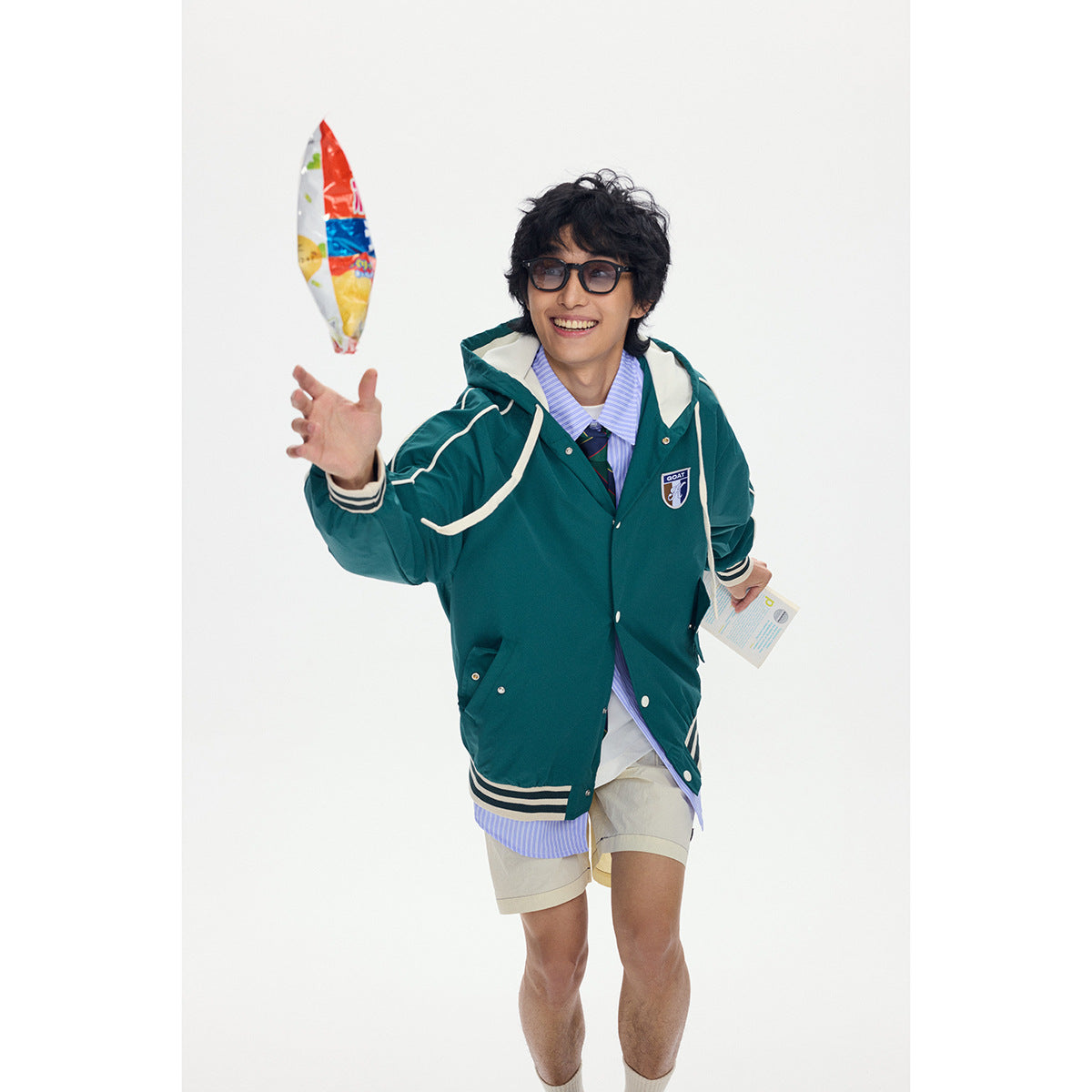 Model in a teal hooded jacket with cream piping and striped trim, tossing a snack against a white background.