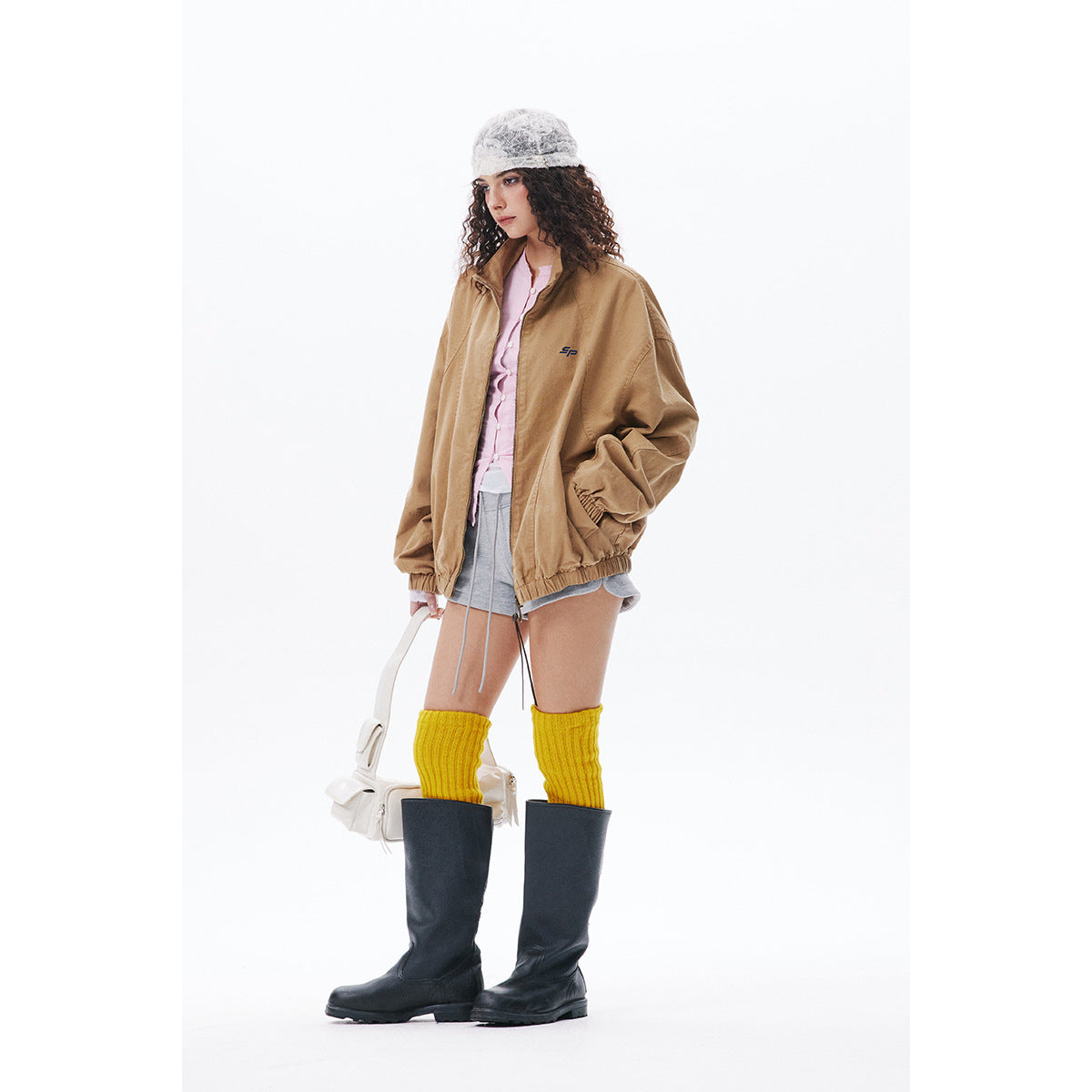 Model in a tan zip jacket worn open over a pink top and gray shorts, with yellow socks and black boots.