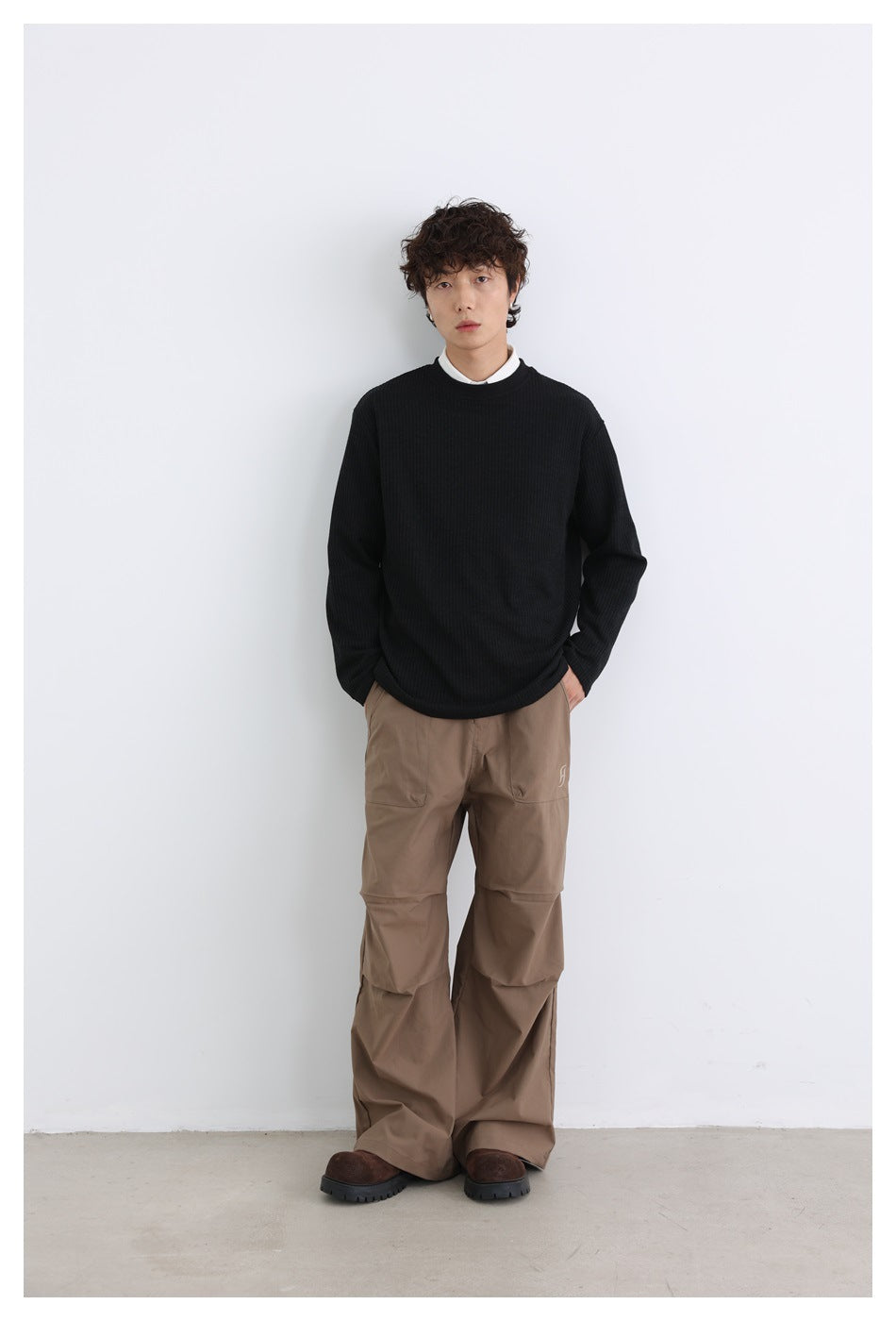 Front view of a model in a black ribbed crewneck over a white collared shirt, with brown trousers.