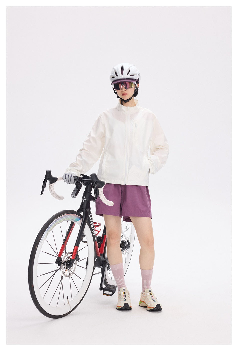 Full-length view of a cyclist wearing a white jacket and plum shorts beside a road bike.