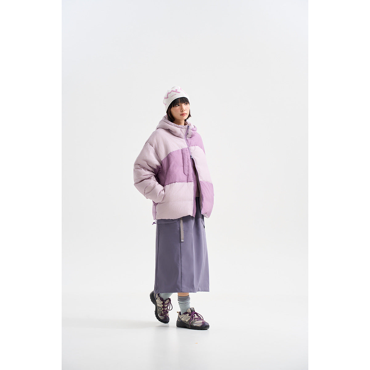 Model wearing a lavender and pale pink color-block hooded puffer jacket with a long gray skirt.