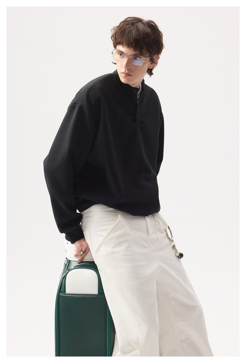 Model in a black long-sleeve button-neck top and white trousers, shown from the thighs up.