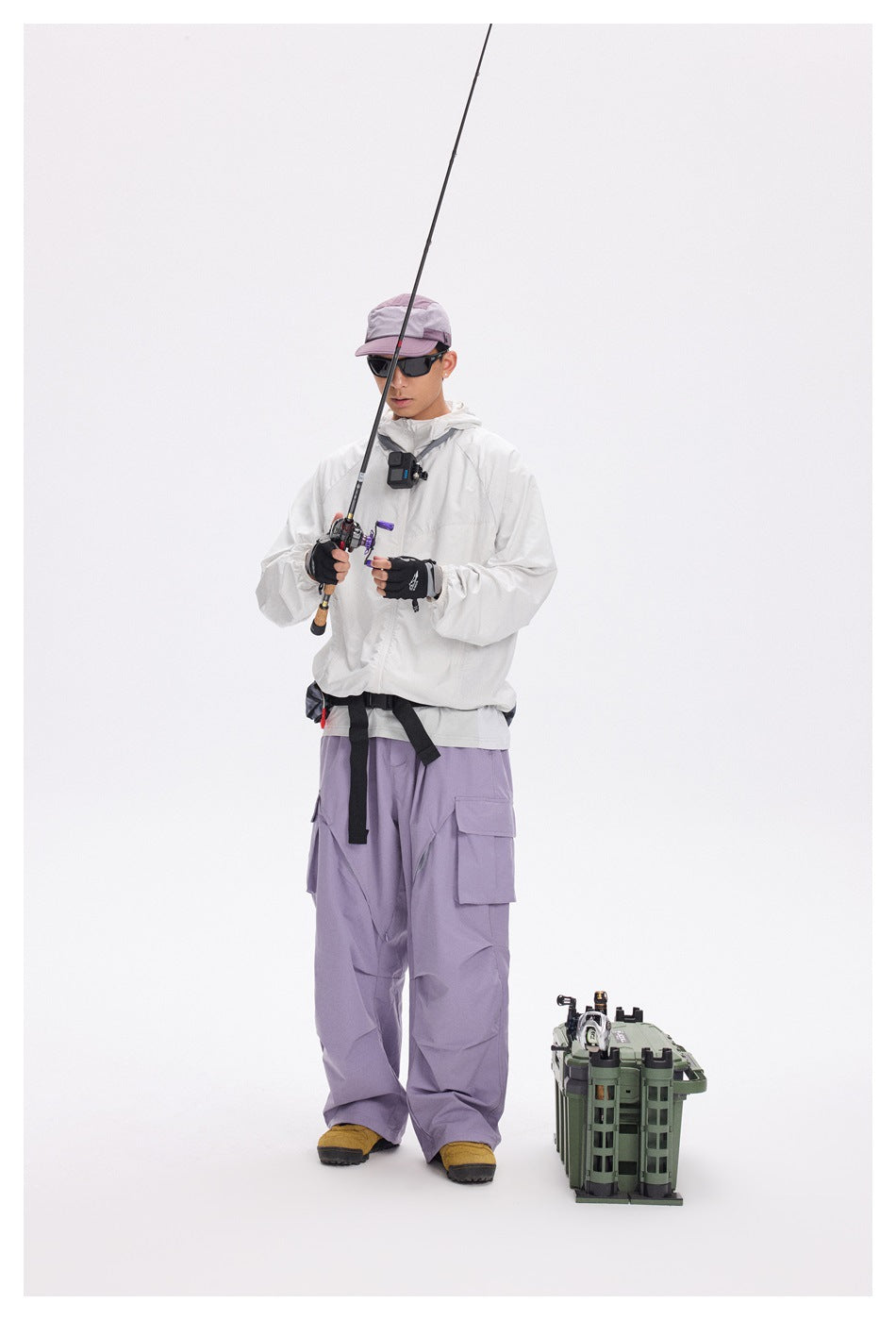 Full-length model wearing a white hooded jacket, lavender cargo pants, and a mauve cap while holding fishing gear.
