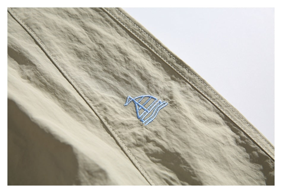 Close-up of beige pants with a small light blue embroidered emblem and stitched seam.