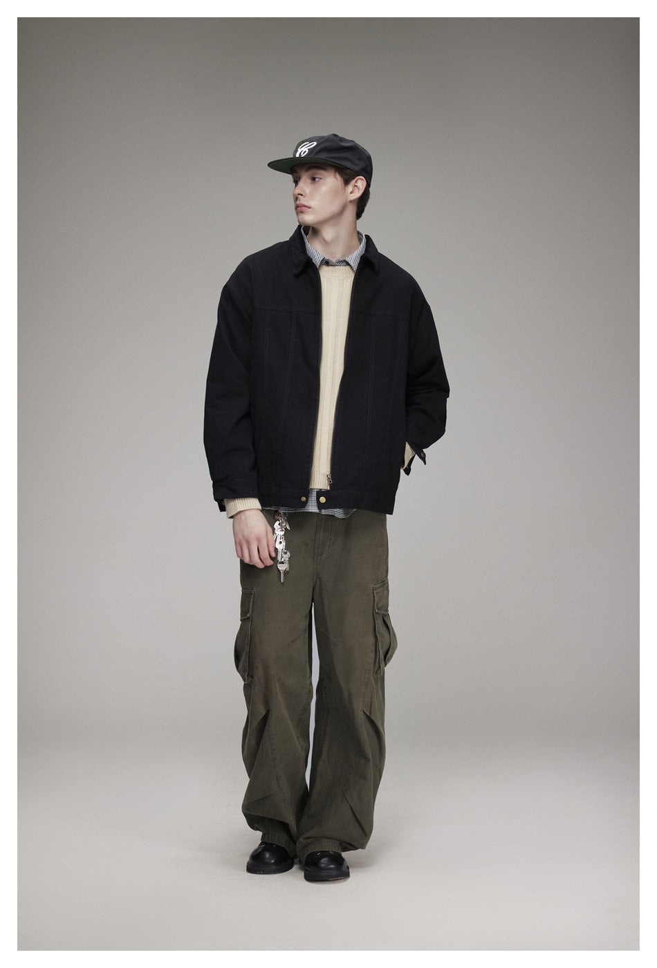 Model wearing black zip jacket over beige sweater with olive cargo pants and cap, front view