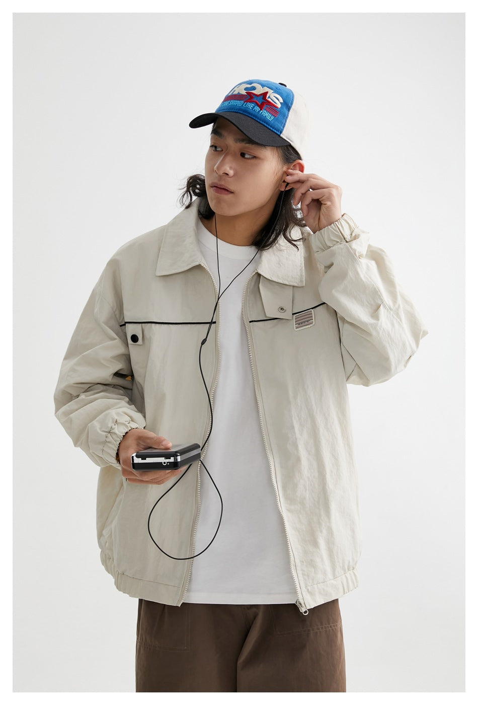 Model wearing an open ivory zip jacket with black chest piping, a white shirt and brown trousers.