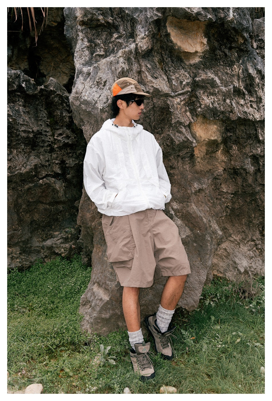 A man leans against rocks wearing a white hooded pullover and taupe cargo shorts with large side pockets.
