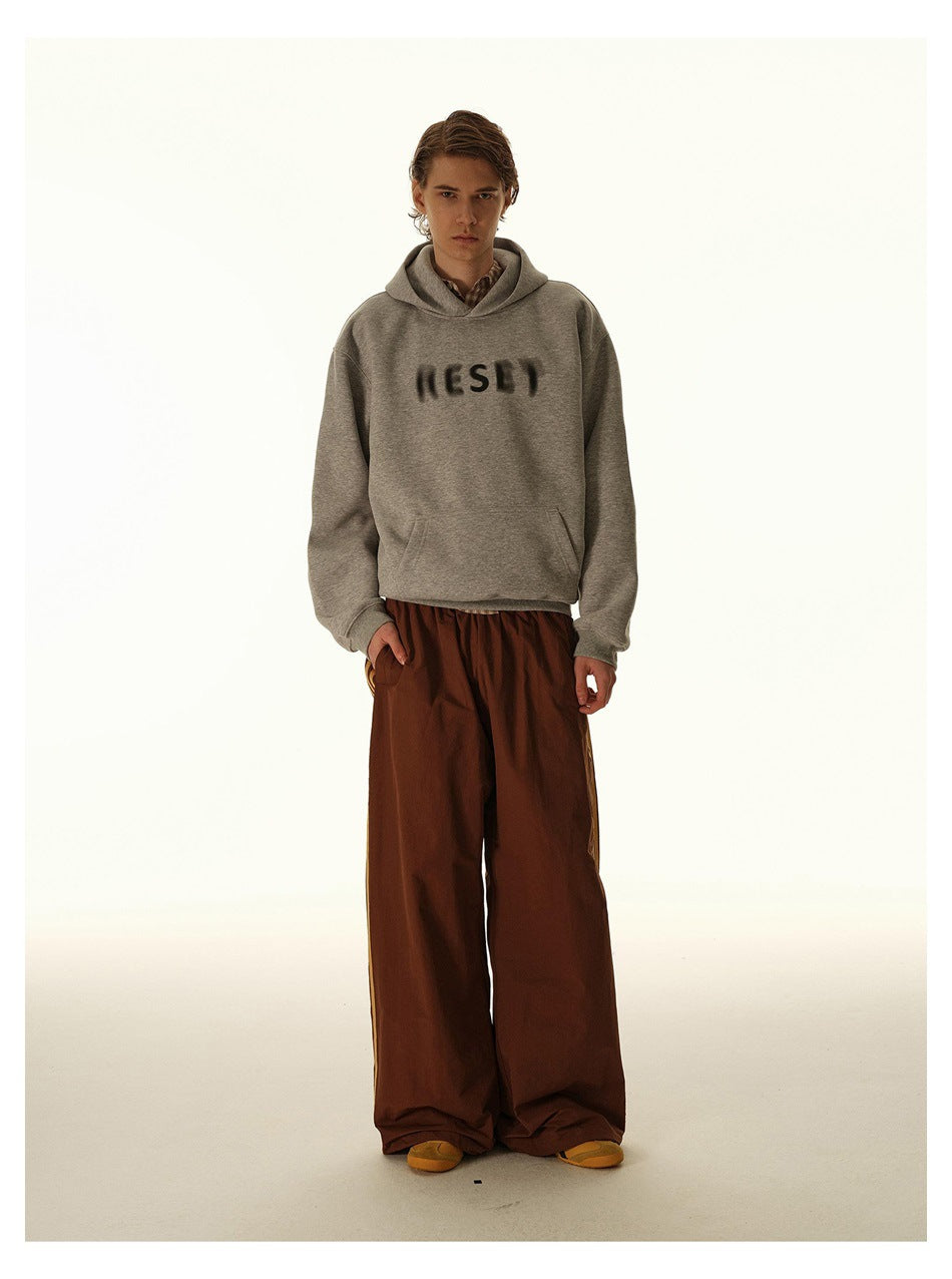 Front view of a model in a light gray hoodie with dark blurred chest lettering and wide rust-brown pants.