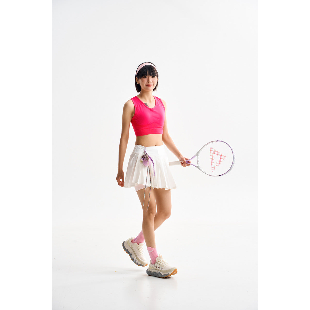 Model wearing a bright pink sleeveless crop top and white pleated tennis skirt, holding a racket.